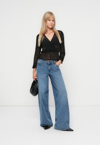 Zwarte blouse met lange mouwen van doorschijnende stof met stippen, gecombineerd met lichtblauwe hoge taille wijduitlopende jeans, aangevuld met een zwarte handtas en puntige schoenen.