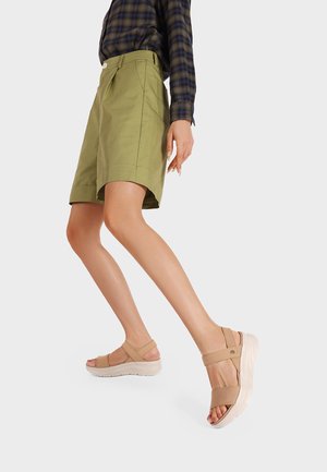 Donna che indossa pantaloncini verde oliva fino al ginocchio e sandali beige con punta aperta, in piedi davanti a uno sfondo chiaro uniforme.