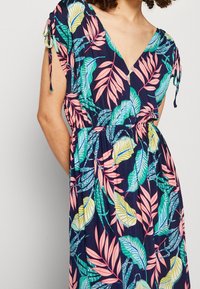 Kobieta ubrana w bezrękawą granatową sukienkę z kolorowymi wzorami tropikalnych liści, z dekoltem w kształcie litery V i związanymi rękawami z wycięciem.