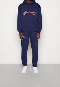 Mörkblå hoodie och sweatpants-set, tillverkat av mjukt tyg. Utrustat med vit och röd "Grimey Mud City" logotyp, med känguruficka och elastiska mudd.