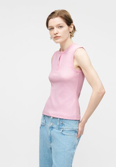 Γυναίκα με κοντά μαλλιά που φοράει χρυσούς κρίκους στα αυτιά, ροζ αμάνικη μπλούζα με λεπτομέρεια κουμπιών και ανοιχτόγαλανά τζιν ψηλόμεσα, κοιτάζει προς την κάμερα.