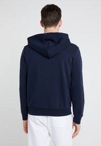 Man i marinblå hoodie och vita byxor som står med ryggen mot kameran mot en ljus, enfärgad bakgrund.