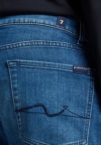 Jeans in denim blu scuro con una tasca posteriore visibile caratterizzata da cuciture uniche. Include un'etichetta con "7" e "for all mankind."
