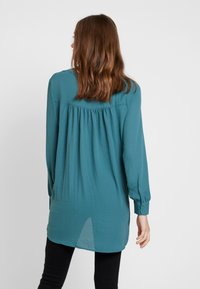 Blusa a maniche lunghe color teal in tessuto leggero, con schiena plissettata e polsini con bottoni, orlo arrotondato e vestibilità morbida.