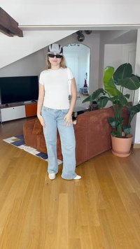 Sieviete baltā T-kreklā un gaiši zilos džinsos, valkājot melnbaltu cepuri un saulesbrilles, stāv gaišā viesistabā ar augu.