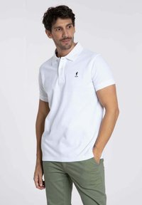 Polo blanc avec un col classique et des manches courtes. Présente un petit logo brodé sur la poitrine. Assorti à un pantalon vert.