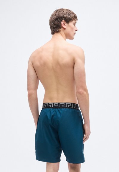 Τιρκουάζ μαγιό σορτς με μαύρη μέση που διαθέτει γεωμετρικό μοτίβο· λεία υφή, σχεδιασμός μέχρι το γόνατο και ελαστική εφαρμογή.