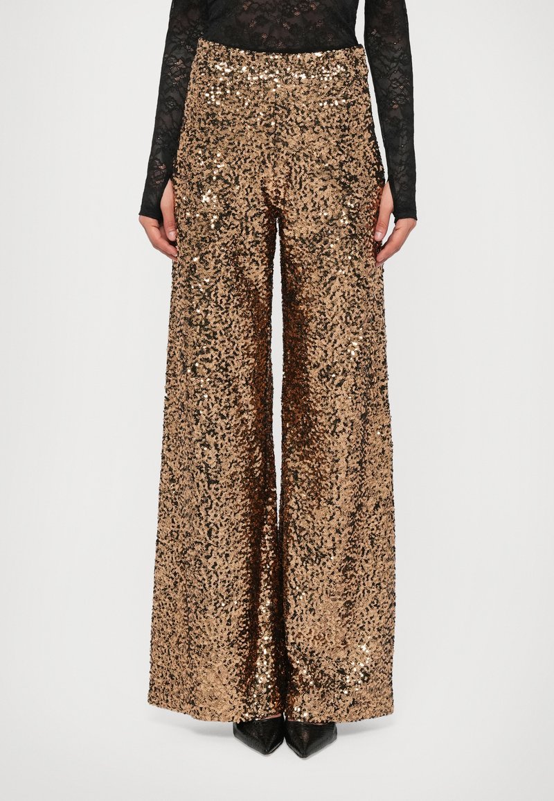 Kuldsetes laialdasetes pükstes, millel on sädelev tekstuur. Omab kõrget vöökohta ja laia lõiget, sobitudes rihmadel mustade kontsakingadega.