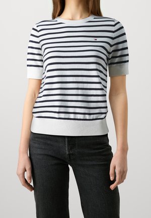 Frau trägt ein weißes kurzärmeliges Oberteil mit marineblauen horizontalen Streifen und dunkle, hoch taillierte Jeans, steht vor einem hellen Hintergrund.