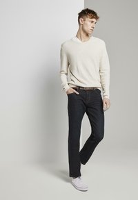 Suéter texturizado de color beige claro llevado sobre una camiseta blanca. Pantalones vaqueros azul oscuro con bolsillos, cinturón marrón y zapatillas blancas. Fondo neutro.