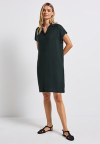 Ciemnozielona sukienka do kolan z kołnierzem i krótkimi rękawami, wykonana z lekkiego materiału. Noszona z czarnymi płaskimi sandałami.