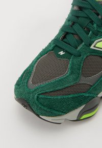 Zöld velúr és hálós sneaker szürke kiegészítőkkel, texturált panelek, párnázott szegély és egy kontrasztos neon zöld logó részlet jellemzi.