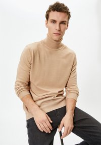 Beige coltrui gemaakt van een zachte stof met geribbelde manchetten en zoom, gecombineerd met donkere spijkerbroeken. Gladde textuur en aansluitend ontwerp.