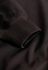 Felpa marrone con polsini a coste e texture liscia, che mette in evidenza i dettagli delle cuciture e il drappeggio morbido del tessuto.