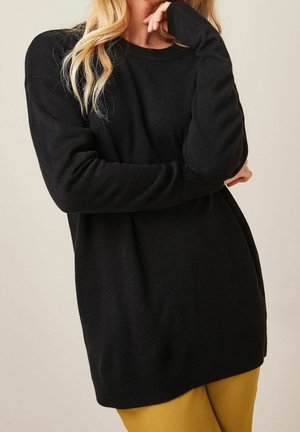 Kobieta z długimi blond włosami, ubraną w czarny sweter i musztardowe spodnie, z założonymi ramionami, ręką blisko brody.