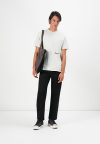 Lys grå t-shirt med logo, sorte lige bukser og en mørkegrå taske. Skoene er marineblå med hvide såler. Enkel, afslappet outfit.