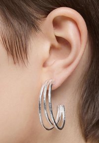 Pendientes de aro plateados con tres lazos entrelazados, que presentan una superficie texturizada y un acabado pulido, llevados en una oreja visible.
