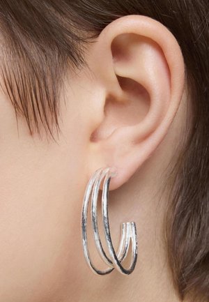 Pendientes de aro plateados con tres lazos entrelazados, que presentan una superficie texturizada y un acabado pulido, llevados en una oreja visible.