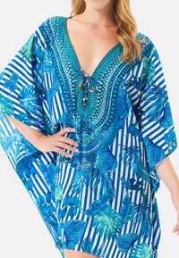Kaftán a rayas azul y blanco con vibrantes patrones de hojas tropicales, escote en V profundo y cordones decorativos en el cuello. Tejido ligero.