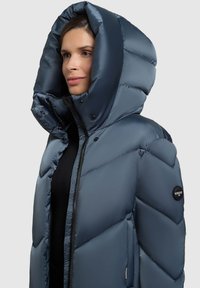 Polsterdatud sinine pufferjakk kõrge krae ja suure kapuutsiga, millel on tõmblukk, trukid ja vasakul varrukal logomärk.