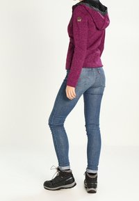 Felpa fucsia con zip e cappuccio nero, tessuto texture e patch con logo, abbinata a jeans skinny blu e scarpe da trekking nere.