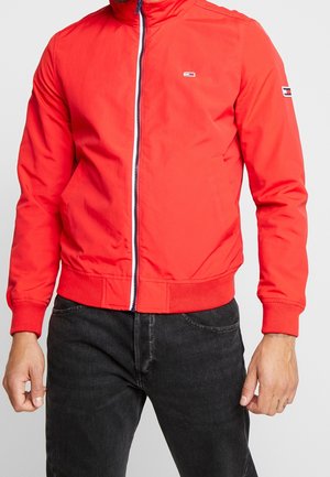 Man draagt een felrode zip-up jas met logo-patches op de borst en mouw, gecombineerd met een zwarte spijkerbroek tegen een effen achtergrond.