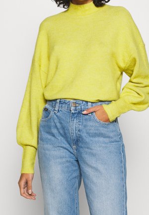 Personne portant un pull à manches longues jaune vif rentré dans un jean taille haute bleu clair, avec une main dans la poche.
