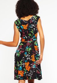 Femme aux cheveux bouclés portant une robe noire sans manches, longueur genou, avec des motifs floraux vifs en orange, bleu, blanc et vert, vue de dos.