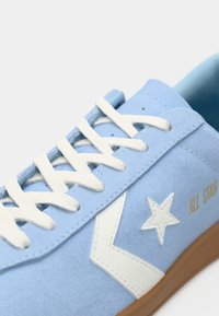 Světle modré semišové tenisky s bílými tkaničkami, které mají bílé logo hvězdy a gumovou podrážku. Zlatý text "ALL STAR" na boku.