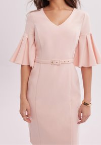 Femme portant une robe cintrée rose pâle avec un col en V, des manches évasées mi-longues, une ceinture assortie, un bracelet en or et une bague en or.
