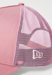 Ružičasta kapa s mrežastim stražnjim dijelom. Ima zaobljeni vrh i izvezeni logo s lijeve strane. Materijal izgleda mekano i teksturirano.