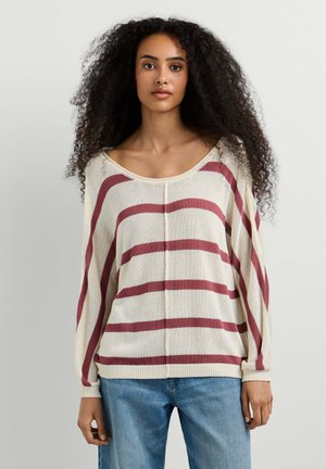 Donna con lunghi capelli ricci che indossa un maglione beige con ampie strisce orizzontali rosse e jeans blu, in piedi davanti a uno sfondo semplice.