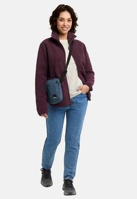 Fleecevest in diep paars, gecombineerd met een licht overhemd en blauwe spijkerbroek. Een klein blauw crossbodytasje maakt de outfit compleet, met zwarte sneakers.
