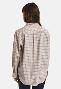Camisa de manga longa em tecido suave bege, com um padrão xadrez em bordô, colarinho abotoado e uma bainha ligeiramente curva nas costas.
