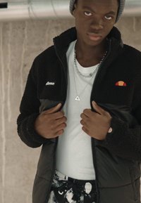Schwarze Fleece- und Nylonjacke mit Reißverschluss, mit einem kleinen Logo. Trägt sie über einem weißen T-Shirt und gemusterten schwarzen Hosen. Silberne Kettenhalskette.
