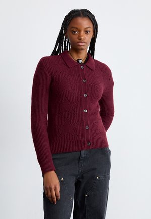 Bordeaux gebreide cardigan, met een kraag, knoopsluiting, een textuurpatroon met sterren en lange mouwen. Gecombineerd met donkere broeken.