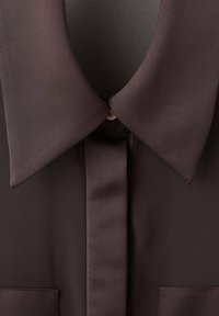 Chemise marron foncé à col avec une patte de boutonnage dissimulée et un petit bouton clair visible près du col.