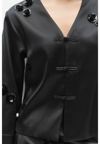 Sort satin-top med V-udskæring, dekoreret med sorte blomsterapplikationer og frøslukning. Glat tekstur med en let glans.