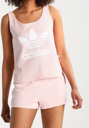 Kobieta nosząca jasnoróżowy bezrękawnik Adidas i pasujące szorty, prezentująca duże logo Adidas na bezrękawniku na jednolitym tle.