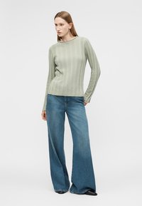 Donna in piedi che indossa una maglia a maniche lunghe verde chiaro a coste e jeans azzurri a gamba larga con scarpe nere, in posa davanti a uno sfondo semplice.
