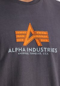 Siva bombažna majica z oranžnim teksturiranim logotipom z zvezdnim vzorcem in napisom "ALPHA INDUSTRIES, KNOXVILLE, TENNESSEE, ZDA."