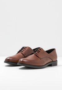 Chaussures habillées en cuir marron avec une texture lisse, un bout arrondi et un design à lacets. Elles présentent un petit talon rectangulaire et des coutures discrètes.