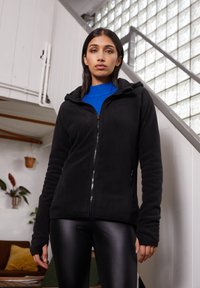 Žena stoji u prostoriji blizu stepenica, nosi crnu jaknu na patentni zatvarač preko plave majice i crne hlače od kože.