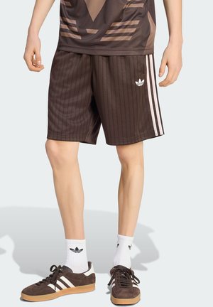 Mężczyzna noszący brązowe spodenki Adidas z czarnymi prążkami i białymi paskami, białe skarpetki Adidas oraz brązowe buty sportowe Adidas z gumowymi podeszwami.