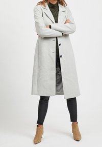 Lys grå uldfrakke med notch-krave, enkeltknappet lukning og to forlommer. Båret over en mørkegrøn bluse og sorte leggings.