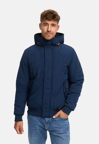 Joven vestido con una chaqueta de invierno azul marino con capucha y jeans azul claro, de pie con una mano en el bolsillo frente a un fondo blanco.