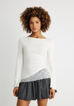 Mladá žena s kučeravými vlasmi má na sebe biele tričko s dlhým rukávom s čipkovým detailom a sivú sukňu s úzkymi prúžkami, stojí pred jednofarebným pozadím.