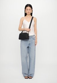Svart lærveske med en strukturert design, som har en bred bunn og en lang justerbar stropp. Modell iført lys topp og blå jeans.