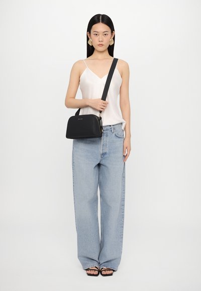 Sac à main en cuir noir au design structuré, avec une large base et une longue sangle réglable. Modèle portant un haut clair et un jean bleu.