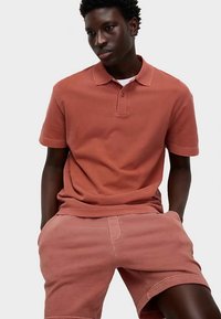 Pikétröja i terrakottafärgad bomullstyg med treknappslås, kombinerad med matchande shorts, med avslappnad passform och mjuk textur.
